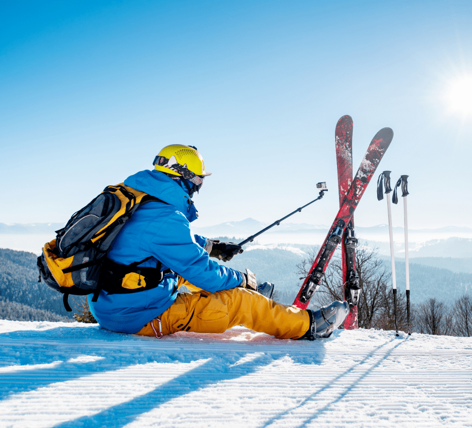 A skier in blue and yellow gear sits on a snowy slope, facing a mountainous landscape, with skis and poles upright beside them.