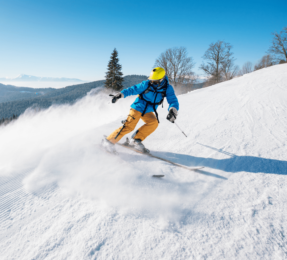 A person in blue jacket and yellow helmet skis down a snowy slope, creating a trail of snow behind. Clear blue sky and trees in the background.