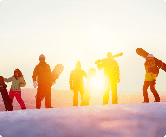 A group of people with snowboards stand on a snowy slope at sunset.
